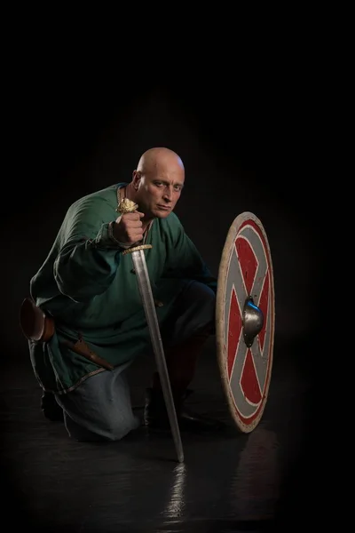 Portrait of a brutal bald man viking in linen clothes with a sword in hands posing against a black background. Early medieval period