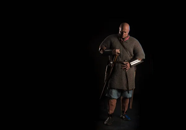 Portrait of a brutal bald-headed Viking in a military mail with a battle ax in his hands posing on a black background. Early medieval period