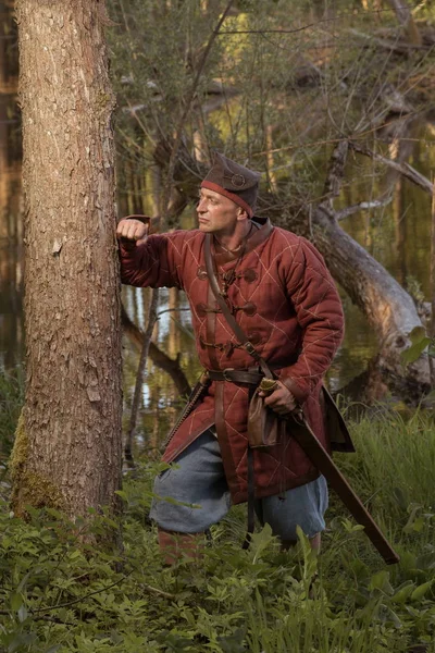Man actor in viking clothes with a sword posing in the forest near lake