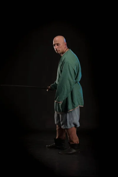 Portrait of a brutal bald man viking in linen clothes with a sword in hands posing against a black background. Early medieval period