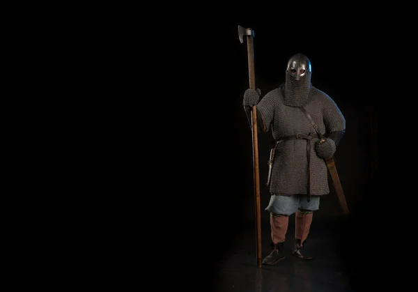 Portrait of a brutal Viking in a military mail with a helmet on his head and with a battle ax in his hands posing on a black background. Early medieval period