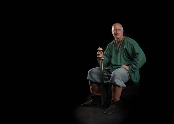 Portrait of a brutal bald man viking in linen clothes with a sword in hands posing against a black background. Early medieval period