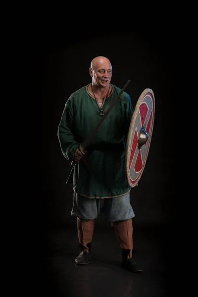 Portrait of a brutal bald man viking in linen clothes with a sword in hands posing against a black background. Early medieval period