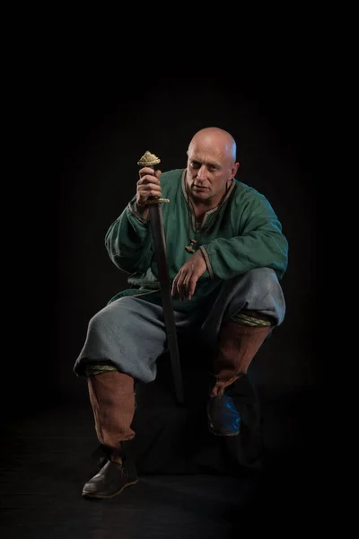 Portrait of a brutal bald man viking in linen clothes with a sword in hands posing against a black background. Early medieval period