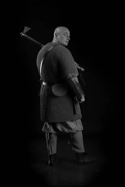 Portrait of a brutal bald man viking in linen clothes with a sword in hands posing against a black background. Early medieval period