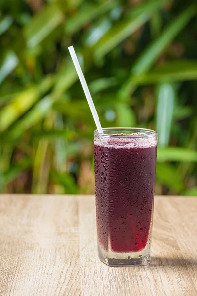 Transparent misted glass with grape juice and straw on background of green bushes