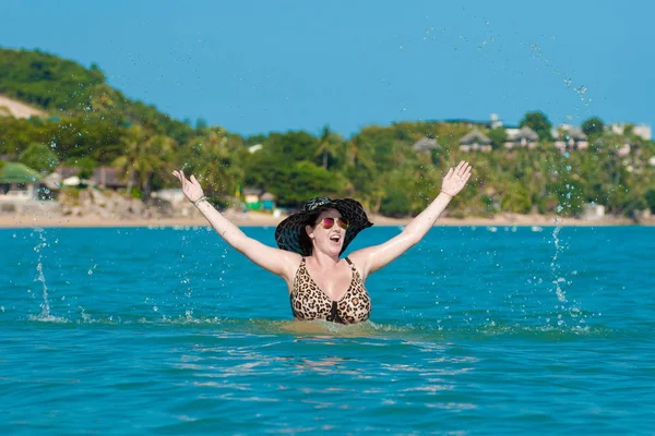 Brunette woman in black hat and sunglasses in blue sea poses for photo