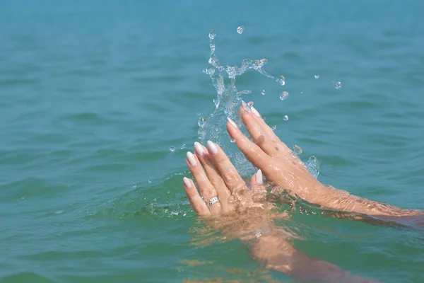 Female hands with white nail polish in the turquoise sea water on the background of the beach