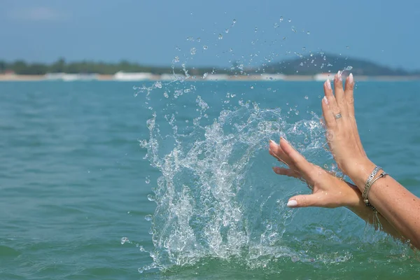 Female hands with white nail polish in the turquoise sea water on the background of the beach