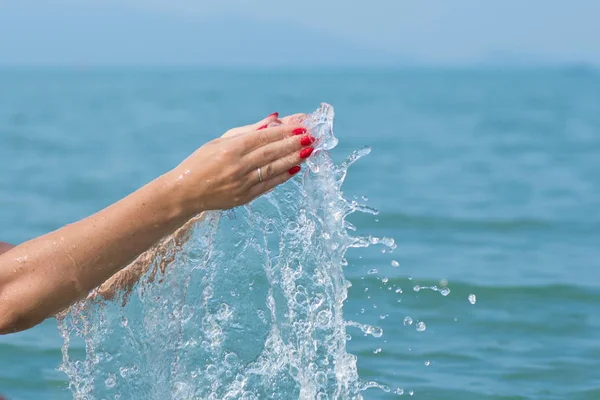 Female hands with white nail polish in the turquoise sea water on the background of the beach