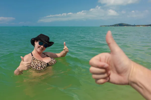 Brunette woman in black hat and sunglasses in blue sea poses for photo with male hand