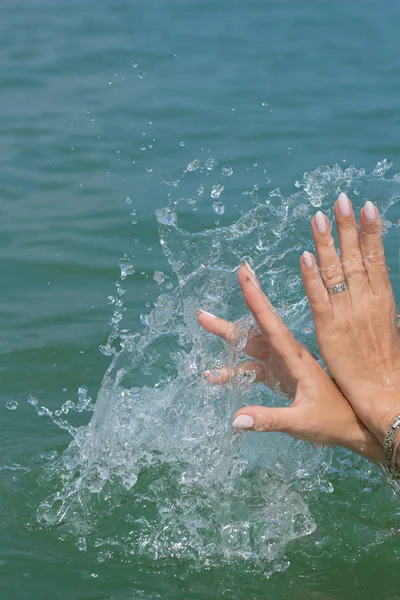 Female hands with white nail polish in the turquoise sea water on the background of the beach