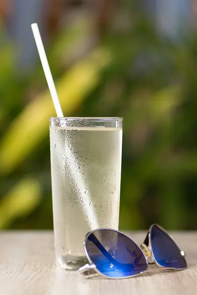 Transparent misted glass with water and straw on table with sunglasses