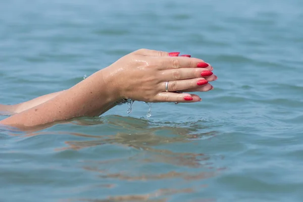 Female hands with white nail polish in the turquoise sea water on the background of the beach