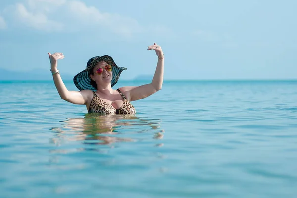 Brunette woman in black hat and sunglasses in blue sea poses for photo