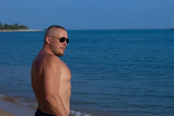 Tanned man with nude torso in sunglasses posing on the beach near the sea at sunset