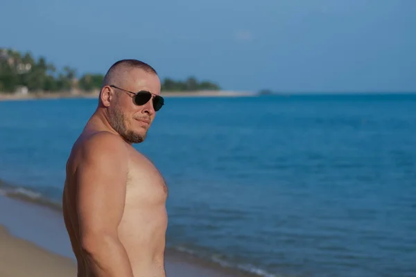 Tanned man with nude torso in sunglasses posing on the beach near the sea at sunset