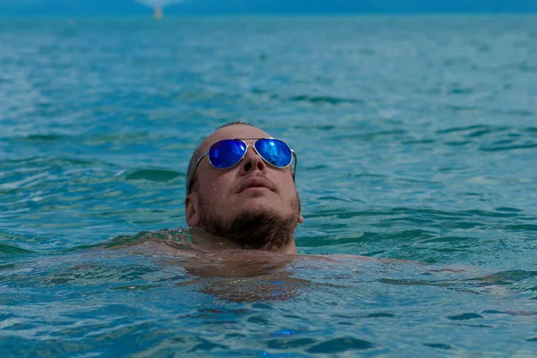 Tanned man with nude torso in sunglasses posing on the beach near the sea at sunset