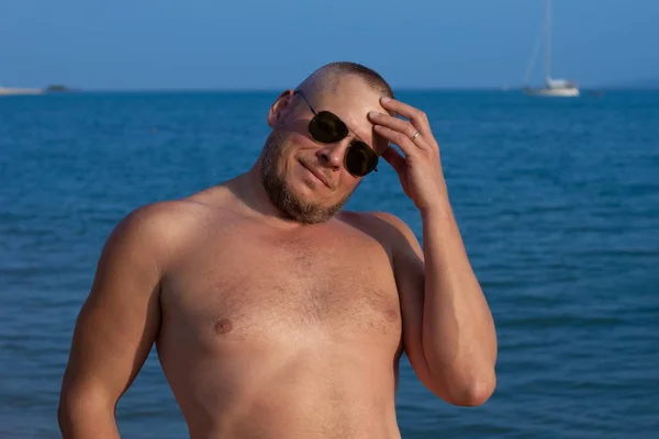 Tanned man with nude torso in sunglasses posing on the beach near the sea at sunset