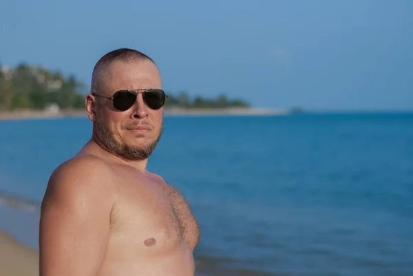 Tanned man with nude torso in sunglasses posing on the beach near the sea at sunset