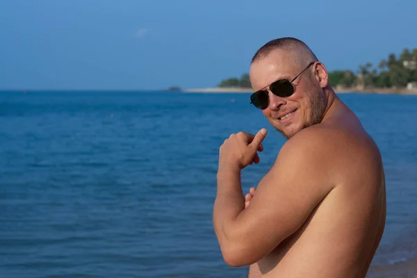 Tanned man with nude torso in sunglasses posing on the beach near the sea at sunset