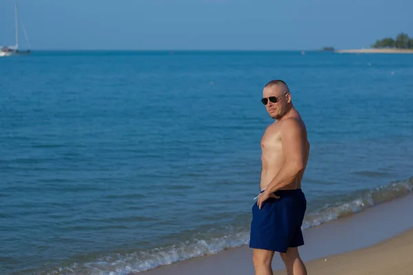 Tanned man with nude torso in sunglasses posing on the beach near the sea at sunset