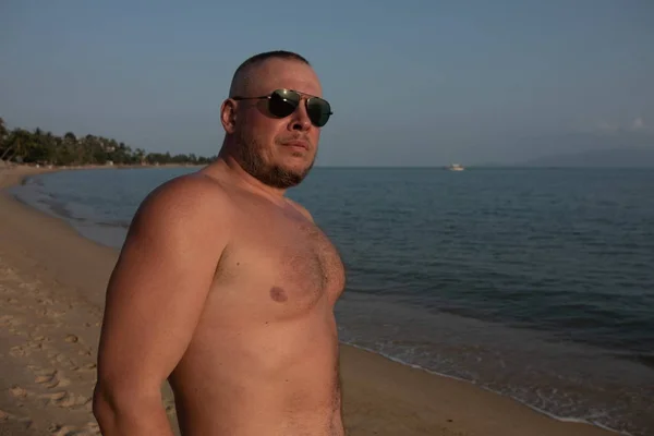 Tanned man  in sunglasses posing on the beach near the sea at sunset