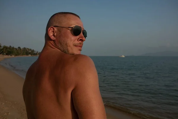 Tanned man  in sunglasses posing on the beach near the sea at sunset