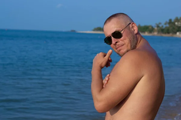 Tanned man with nude torso in sunglasses posing on the beach near the sea at sunset