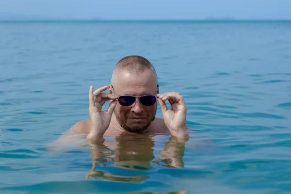 Tanned man with nude torso in sunglasses posing on the beach near the sea at sunset