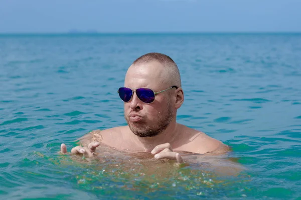 Tanned man with nude torso in sunglasses posing on the beach near the sea at sunset