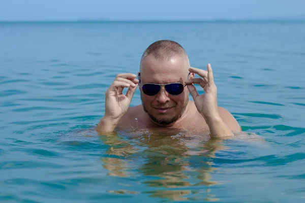 Tanned man with nude torso in sunglasses posing on the beach near the sea at sunset