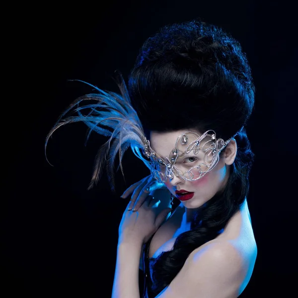 brunette woman with high hair, a mask with feathers and corset in old style on a black background