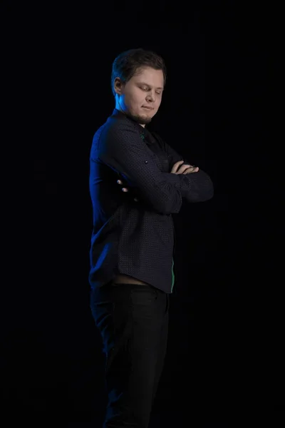 Young blond man in black clothes playing on a musical synthesizer and posing on a black background with blue light