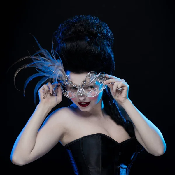 brunette woman with high hair, a mask with feathers and corset in old style on a black background