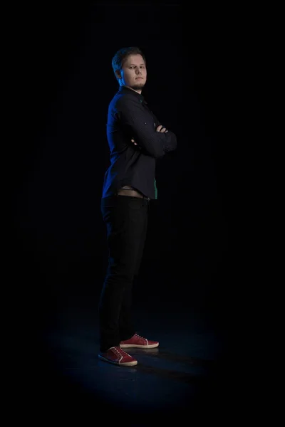 Young blond man in black clothes playing on a musical synthesizer and posing on a black background with blue light