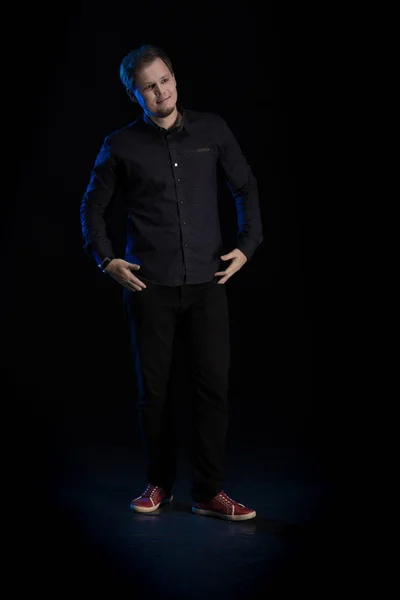 Young blond man in black clothes playing on a musical synthesizer and posing on a black background with blue light