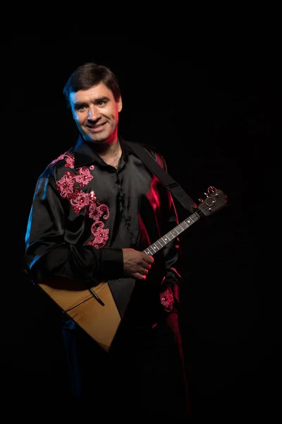 brunette man in a folk shirt plays a balalaika in scenic blue and red light on a black stage