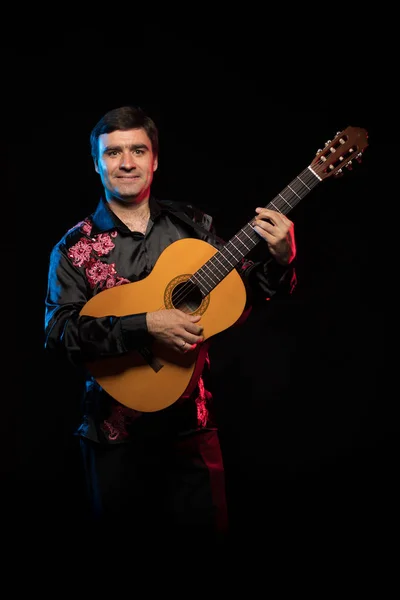 dark-haired man in black shirt playing guitar in blue and red scenic light on dark stage