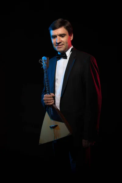 Artist musician, dark-haired man in black suit and bow tie, playing balalaika in blue and red scenic light on dark stage