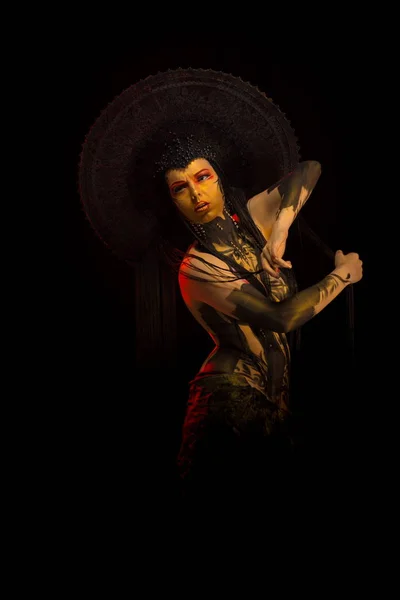 Actress woman in black headdress, body painting, golden figure on body and face, posing on black background