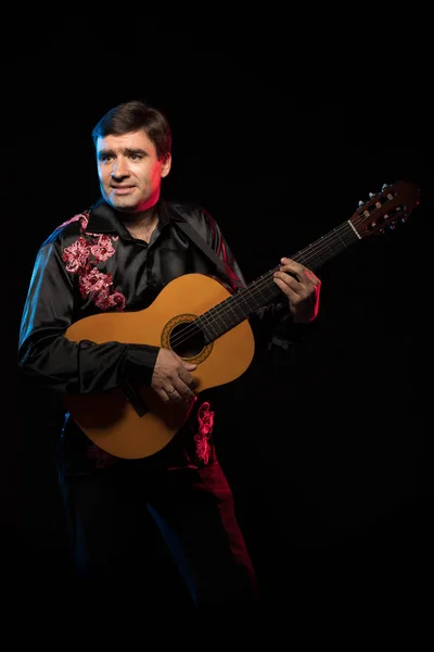 dark-haired man in black shirt playing guitar in blue and red scenic light on dark stage