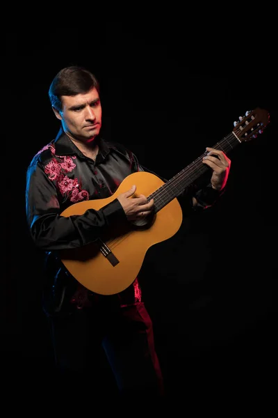 dark-haired man in black shirt playing guitar in blue and red scenic light on dark stage