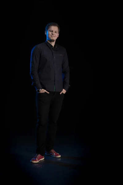 Young blond man in black clothes playing on a musical synthesizer and posing on a black background with blue light