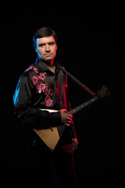 brunette man in folk shirt playing balalaika in scenic blue and red light on black stage