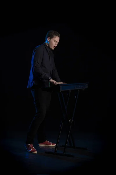 Young blond man in black clothes playing on a musical synthesizer and posing on a black background with blue light