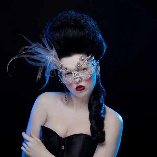 brunette woman with high hair, a mask with feathers and corset in old style on a black background