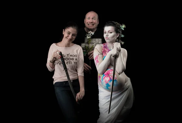 pregnant woman with bodypainting and swords posing on black background with man and woman