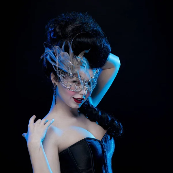 brunette woman with high hair, a mask with feathers and corset in old style on a black background