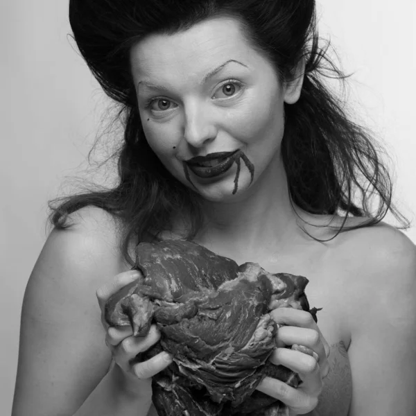 emotional brunette actress in the role of a vampire with blood drops on his face and a piece of bloody meat. woman in a transparent lace dress and lush hair with white feathers on a white background